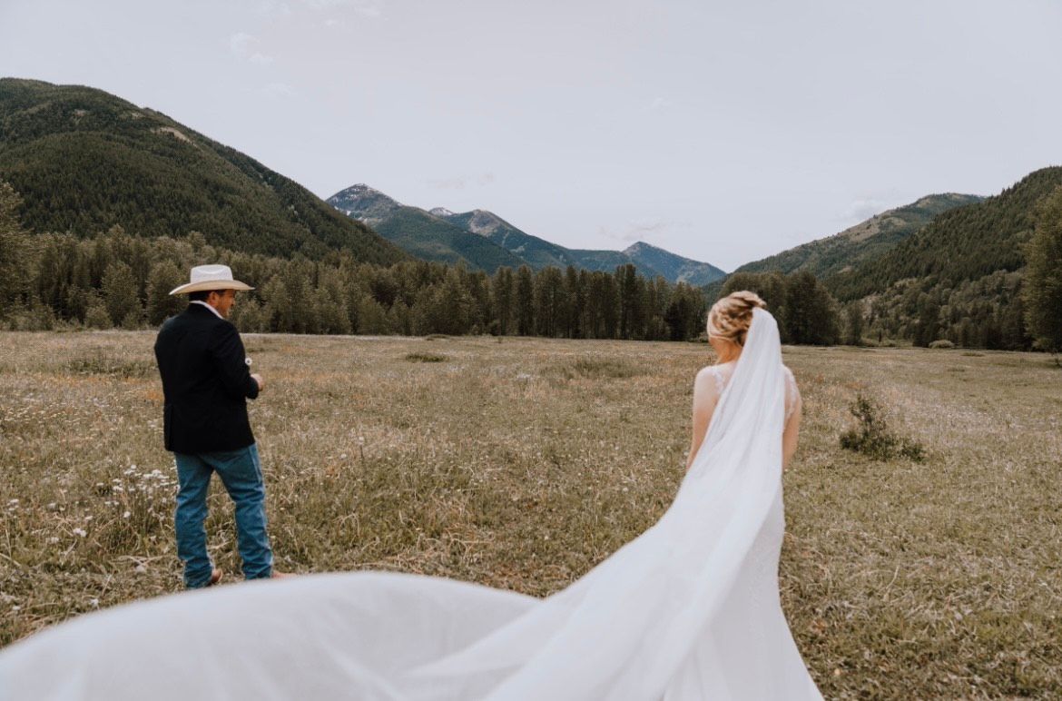 A bride and groom are standing in a field with mountains in the background.
