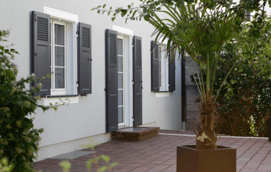 Vor einem Haus mit Fensterläden steht eine große Topfpflanze.