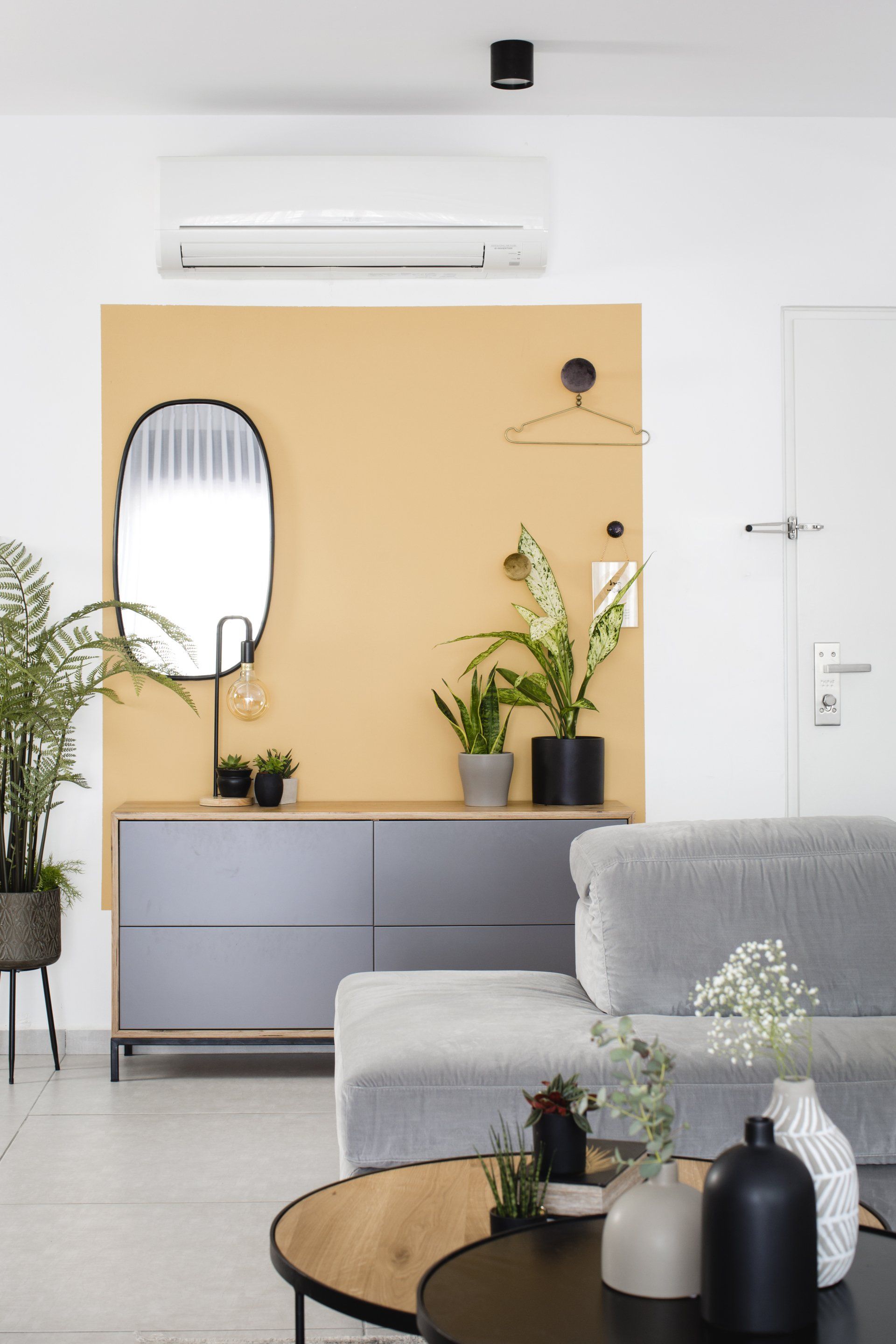 צביעת חלק מקיר בצבע בולט מוסיף עניין לחלל הלבשת חלל סלון ומטבח בגווני אפור שחור וחרדל. שפע של צמחים.  שחור-אפור וכל מה שבניהם בעיצוב של מעצבת פנים ומלבישה בתים ג׳ני ויאליך jenyvstudio