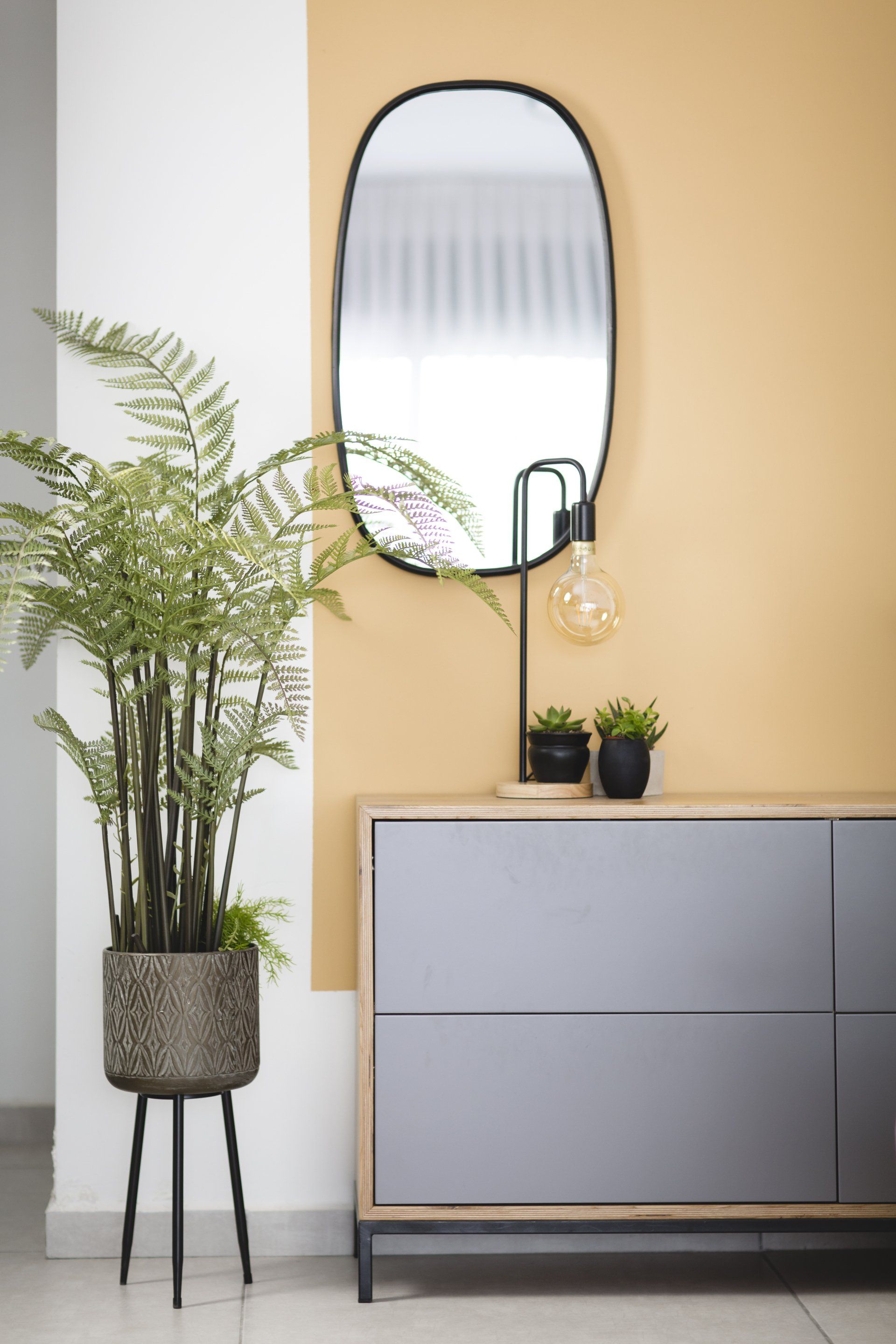 צביעת חלק מקיר בצבע בולט מוסיף עניין לחלל הלבשת חלל סלון ומטבח בגווני אפור שחור וחרדל. שפע של צמחים.  שחור-אפור וכל מה שבניהם בעיצוב של מעצבת פנים ומלבישה בתים ג׳ני ויאליך jenyvstudio