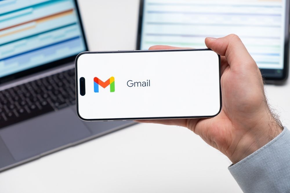 Hand holding a smartphone with Gmail logo in front of a laptop screen