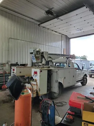 A white utility truck with a crane parked inside a workshop, with a large orange gas cylinder in the foreground.