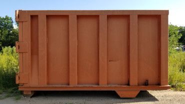 A large, empty orange metal dumpster sits outdoors on a gravel surface against a backdrop of trees.