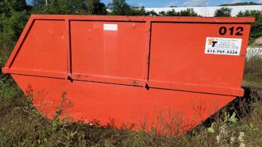 An orange open-top dumpster sits in a grassy, overgrown field with a white logo and the number 012 printed on its side.