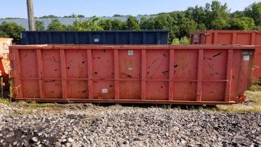 A large, weathered red roll-off dumpster sitting on a gravel lot in front of a blue dumpster and green trees.