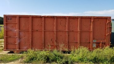 A large, orange, rectangular industrial dumpster sits outdoors on grass under a clear blue sky.