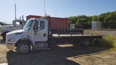 A white flatbed truck with a red container on the back parked on a dirt lot next to another white truck.