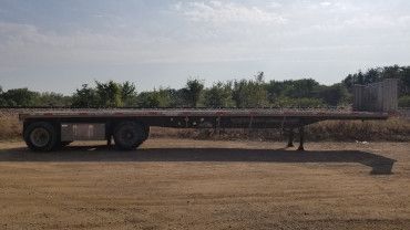 A flatbed semi-trailer parked on a dirt lot, featuring a bulkhead at the front and wheels at the rear.