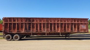 A long, rusted brown industrial trailer with two sets of rear wheels parked on a dirt lot under a clear blue sky.
