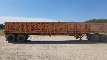 A side view of a large, weathered, orange-brown rectangular trailer resting on a dirt lot under a clear blue sky.