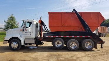 A side view of a white roll-off truck hauling a large orange waste container on a dirt lot under a blue sky.