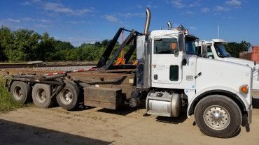 A white roll-off dump truck parked on a dirt lot against a clear blue sky.