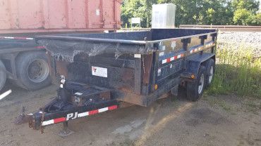 A black PJ dump trailer parked on a gravel lot next to a large red shipping container.