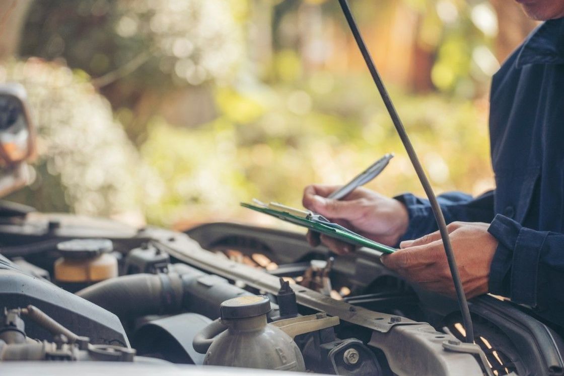 Mechanic inspecting car engine, writing on clipboard, outdoors.