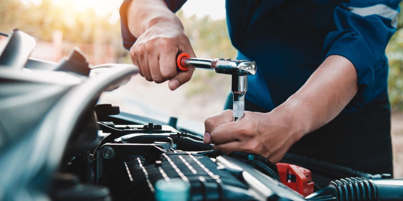 Mechanic working on a car engine with a wrench. The person wears a blue jumpsuit. Sunlight in the background.