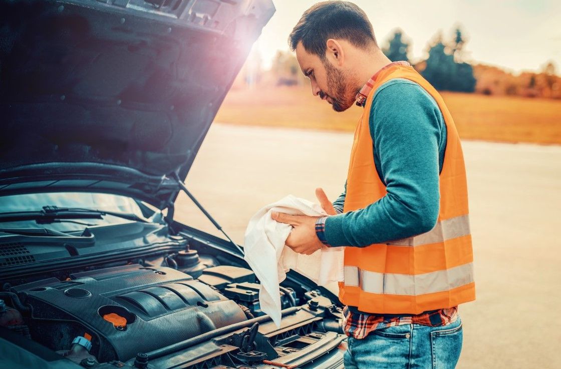 Man in an orange vest inspecting engine of broken-down car, outdoors.