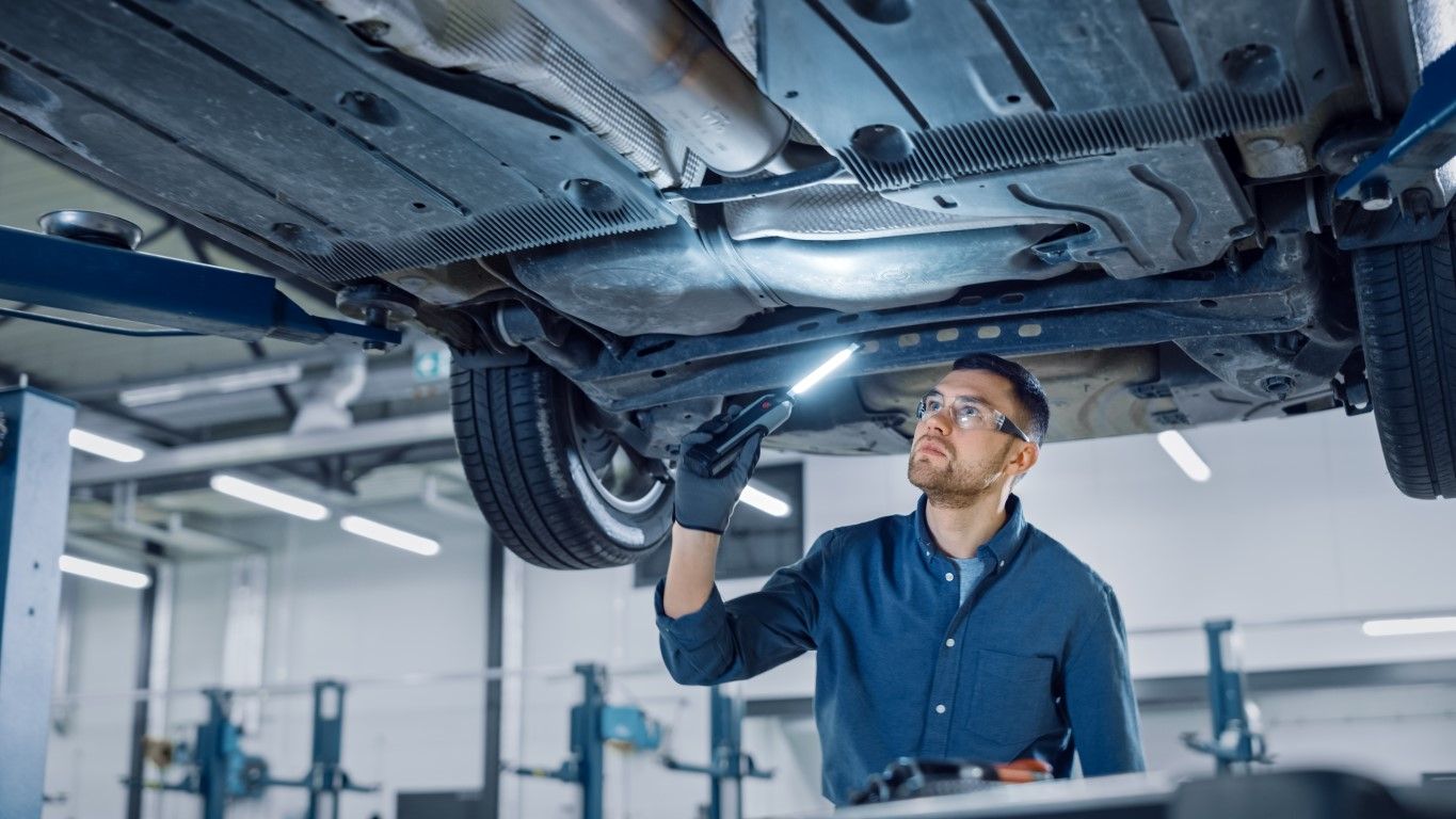 Mechanic in blue shirt examining undercarriage of a car with a flashlight in a well-lit garage.