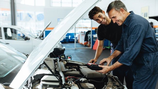Two mechanics examine a car engine in a garage. One older, one younger, smiling.