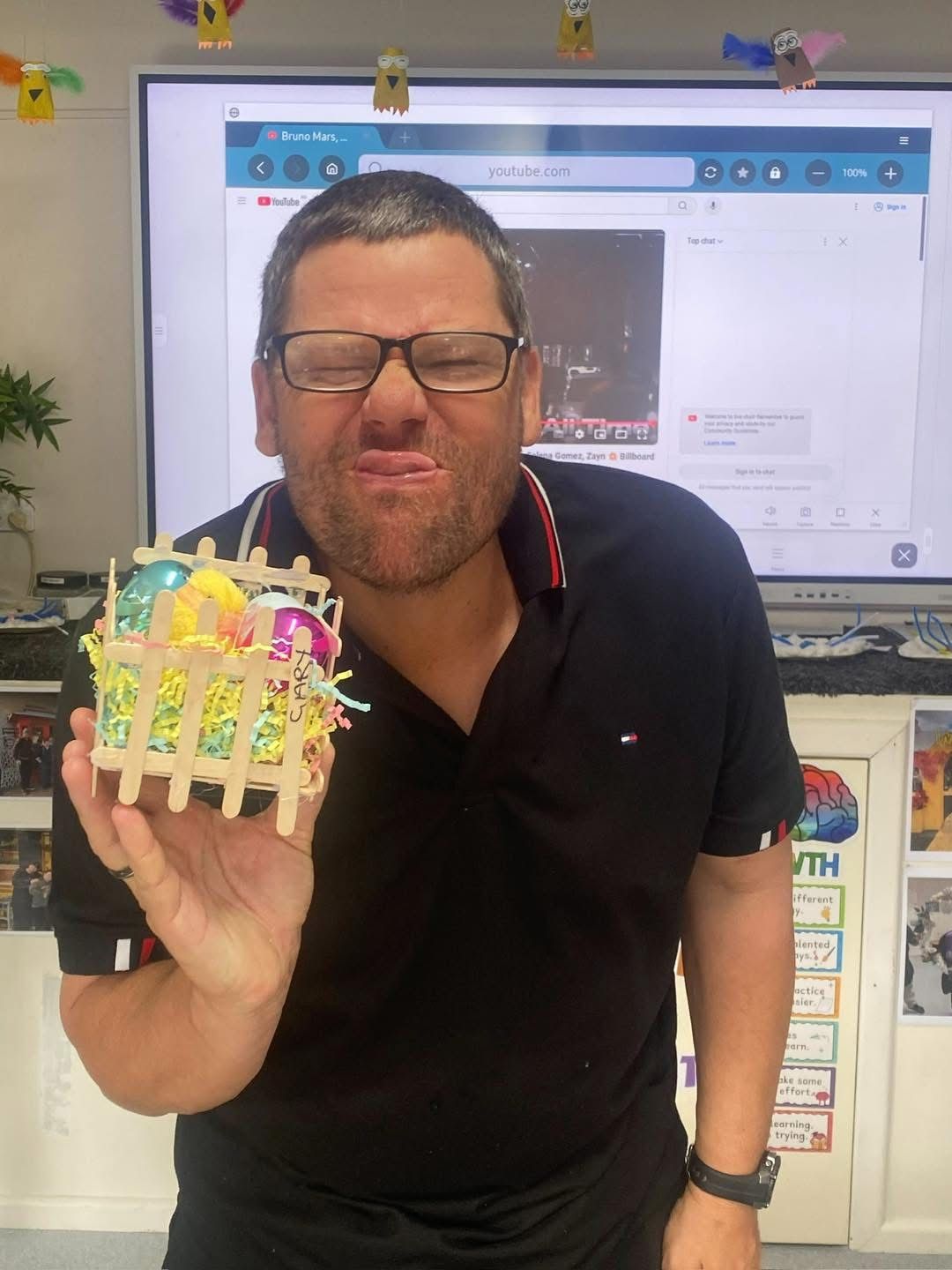 Daily activities. A man wearing glasses is holding a small easter basket in front of a large screen.