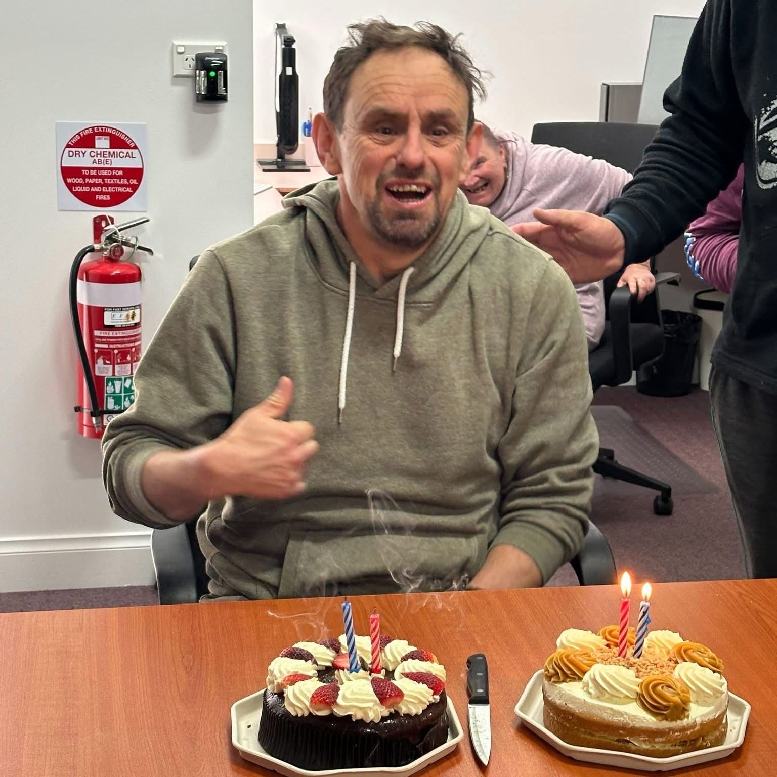 A man is sitting at a table with two cakes and giving a thumbs up