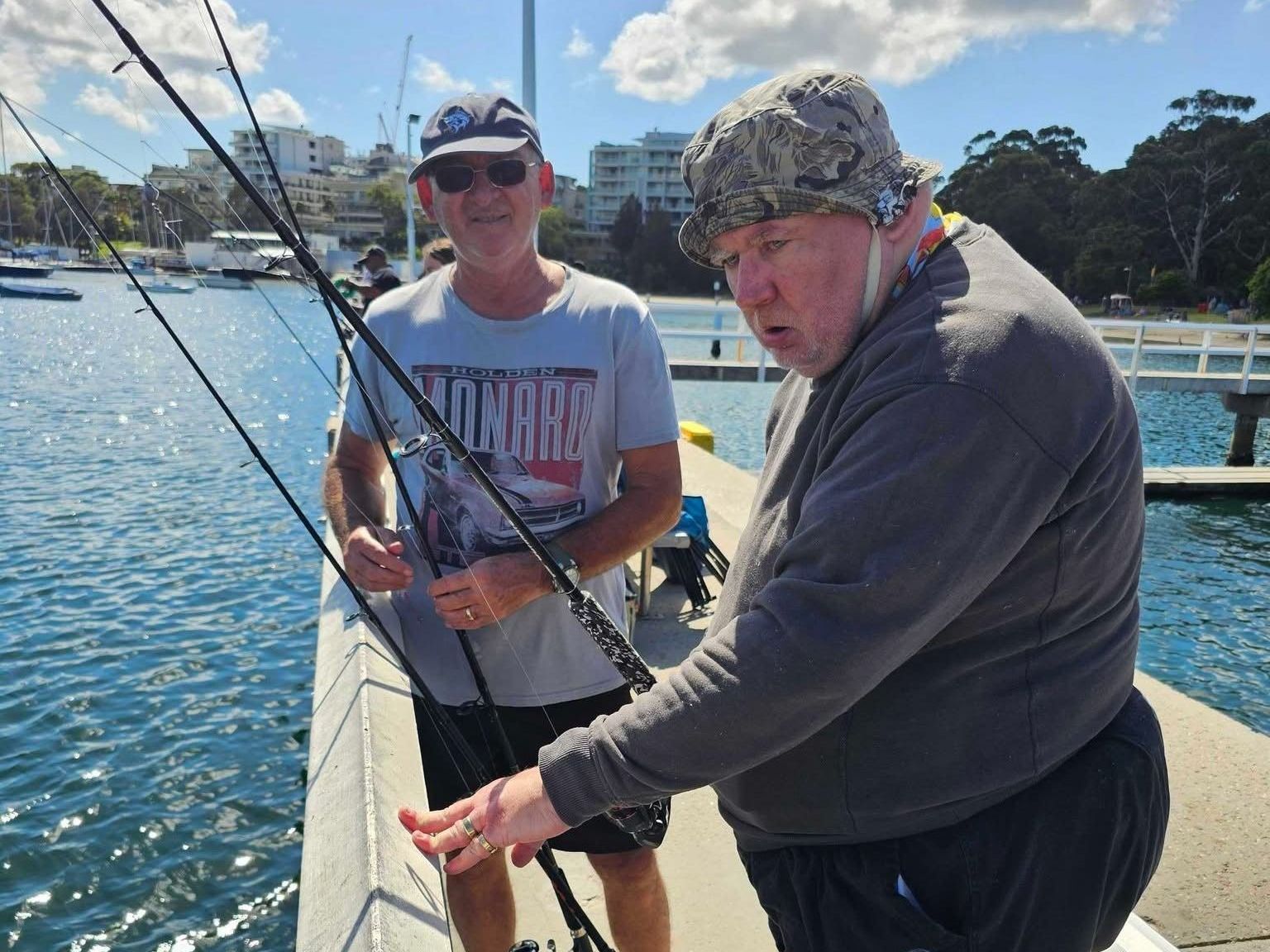 Support worker with client on fishing outing. Two men are fishing on a boat and one of them is wearing a shirt that says always