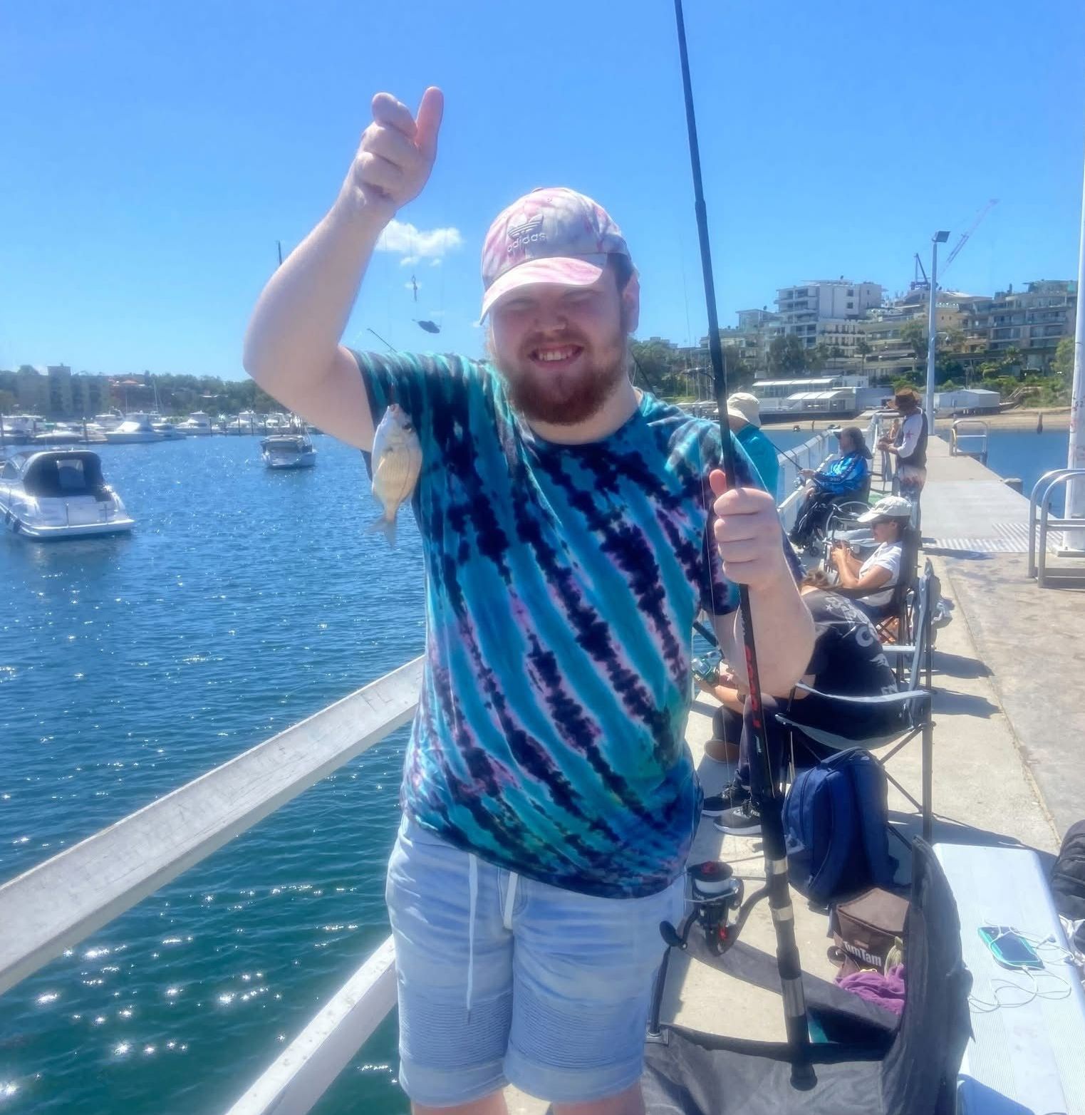Social fishing group. A man in a tie dye shirt is holding a fishing rod and giving a thumbs up