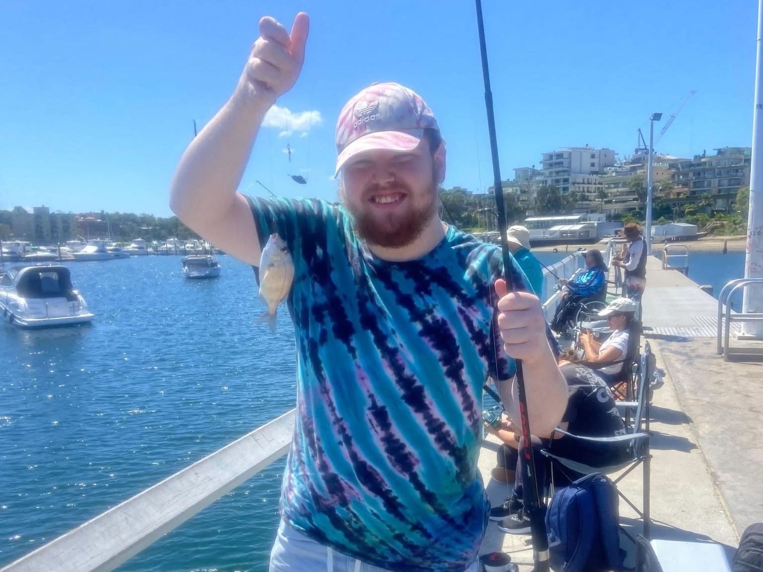 Fishing group outings. A man in a tie dye shirt is holding a fishing rod and a fish