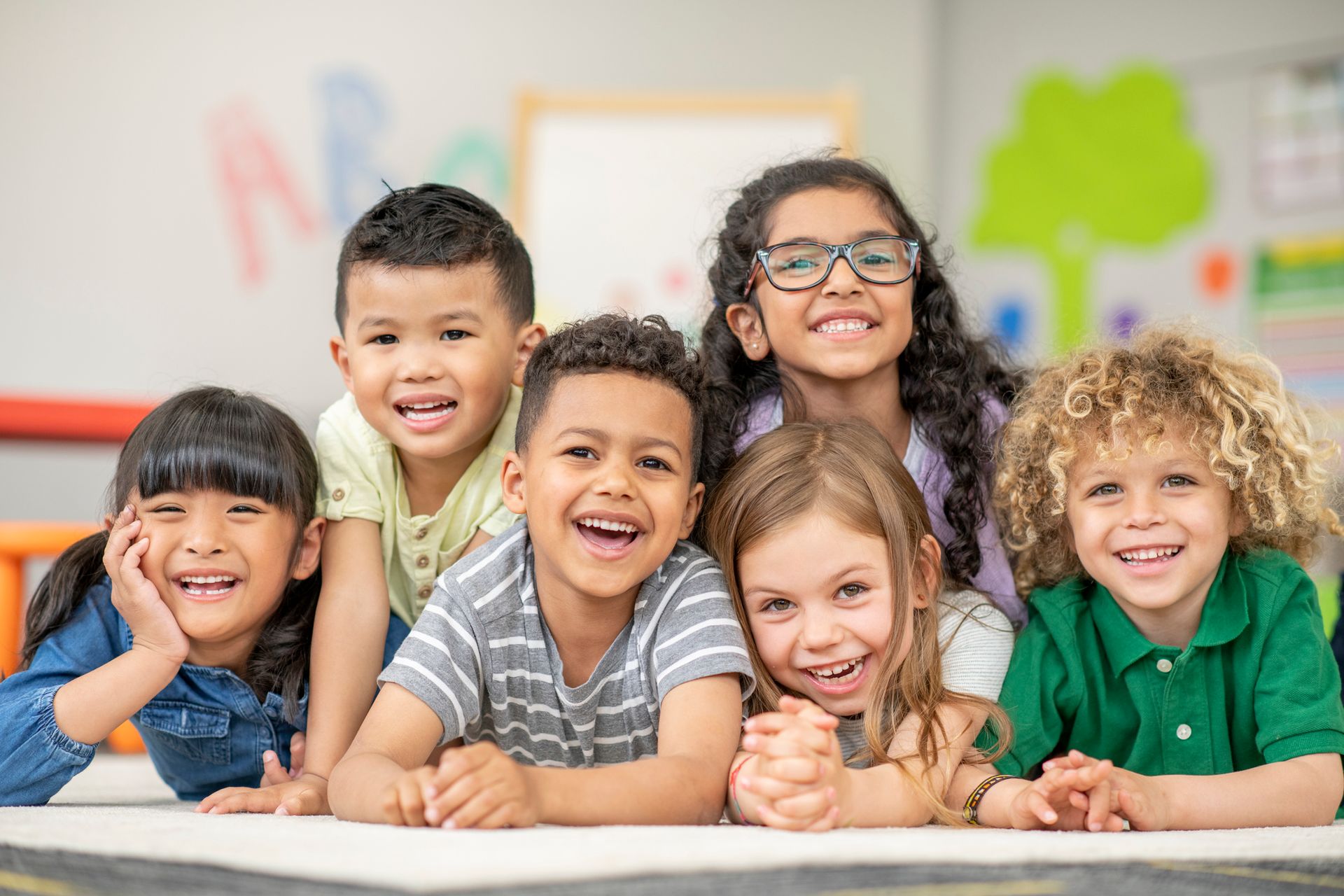 Six diverse children smiling, lying on a rug in a classroom.