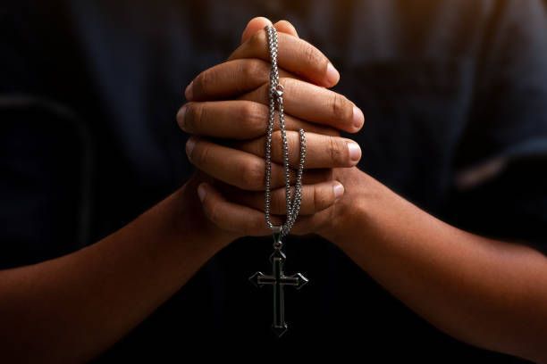 Hands clasped in prayer, holding a rosary with a crucifix. Dark background.