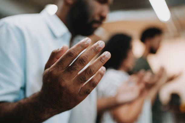 Man with hands raised, praying in a group indoors. Focus on the hands, blurred background.