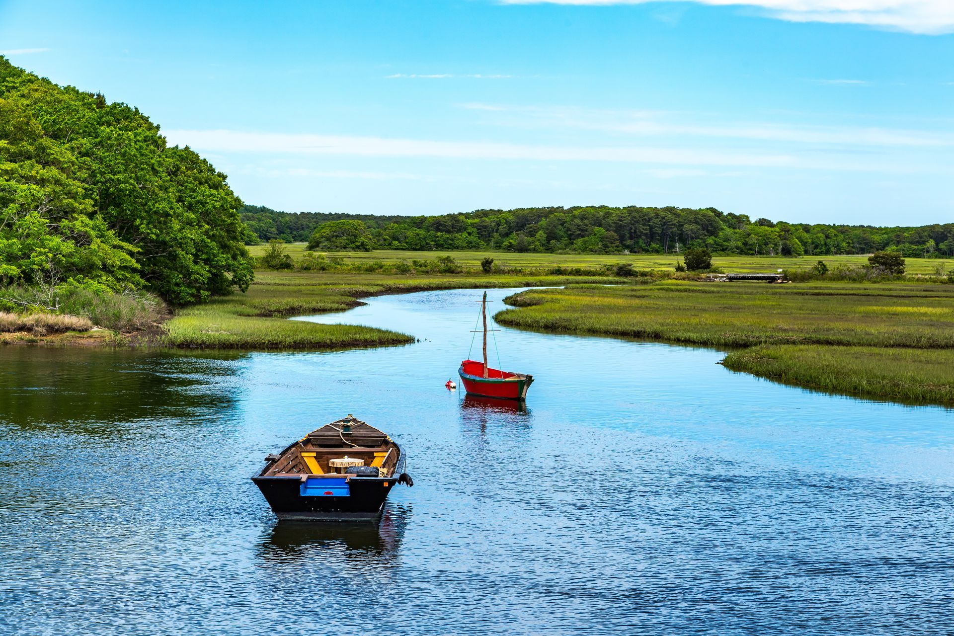Two boats on a calm river, red sailboat and black rowboat, surrounded by green marsh and trees under blue sky.