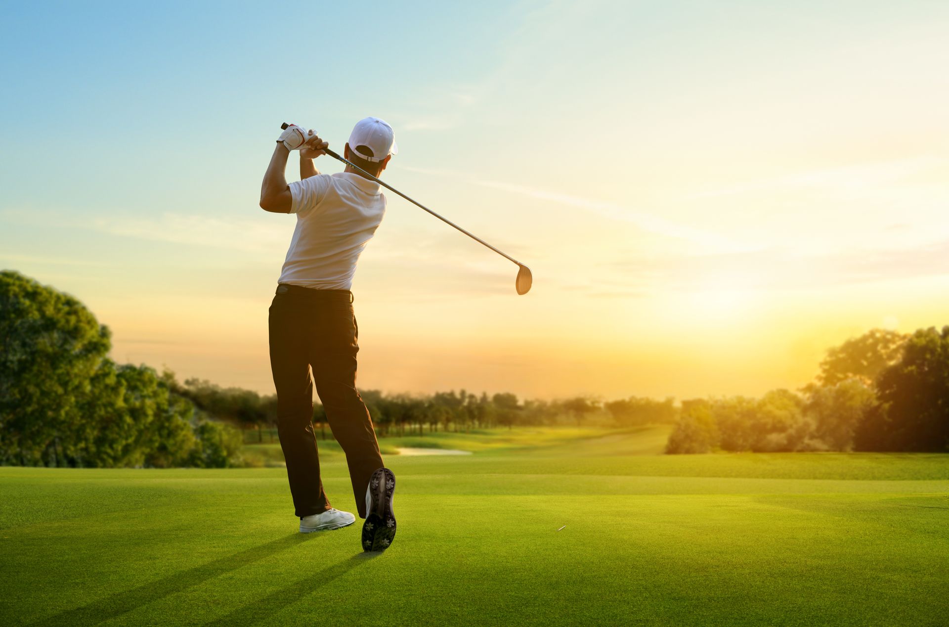 A man is swinging a golf club on a golf course at sunset.