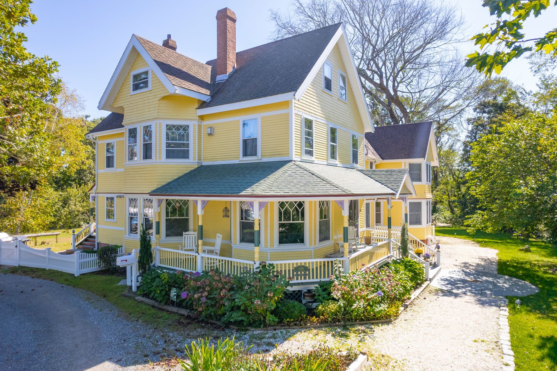Yellow Victorian house with a wraparound porch, set amidst trees and a gravel driveway.