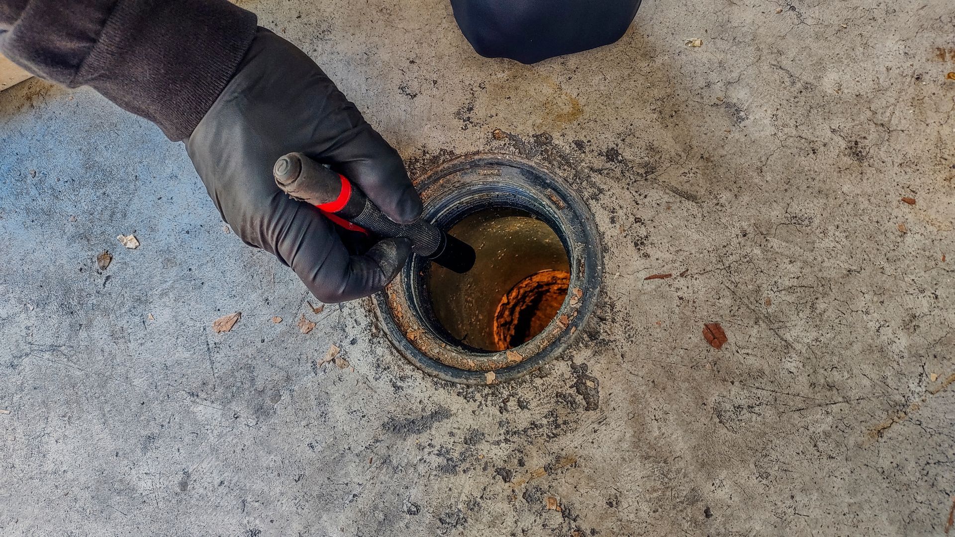 Gloved hand using red-handled tool to inspect a circular drain opening in concrete floor