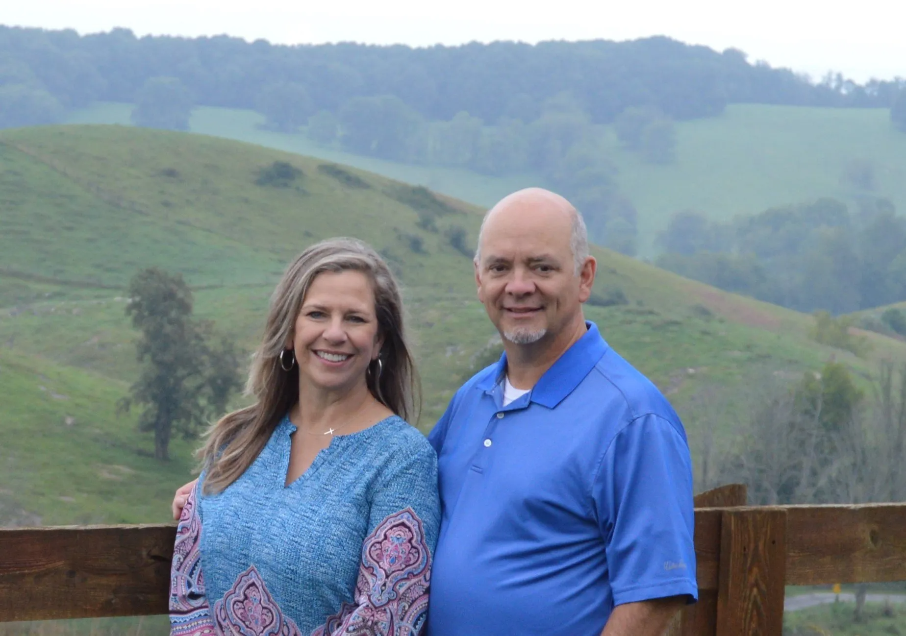 Couple poses with green hills in the background. Woman in blue patterned shirt, man in blue polo.