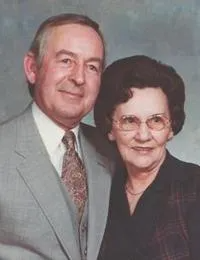 An older man and woman posing together. The man wears a suit and tie. The woman wears glasses and a blazer.