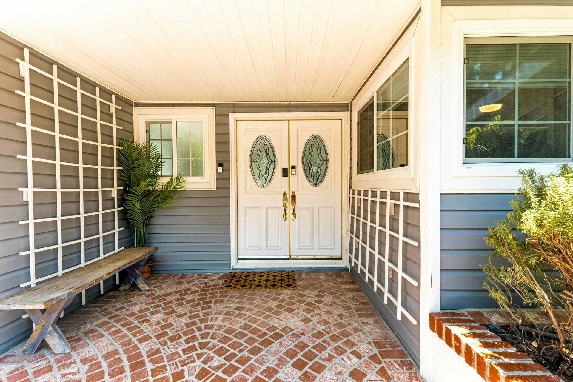 Front porch with white double doors, brick floor, bench, and potted plants under a covered entry.