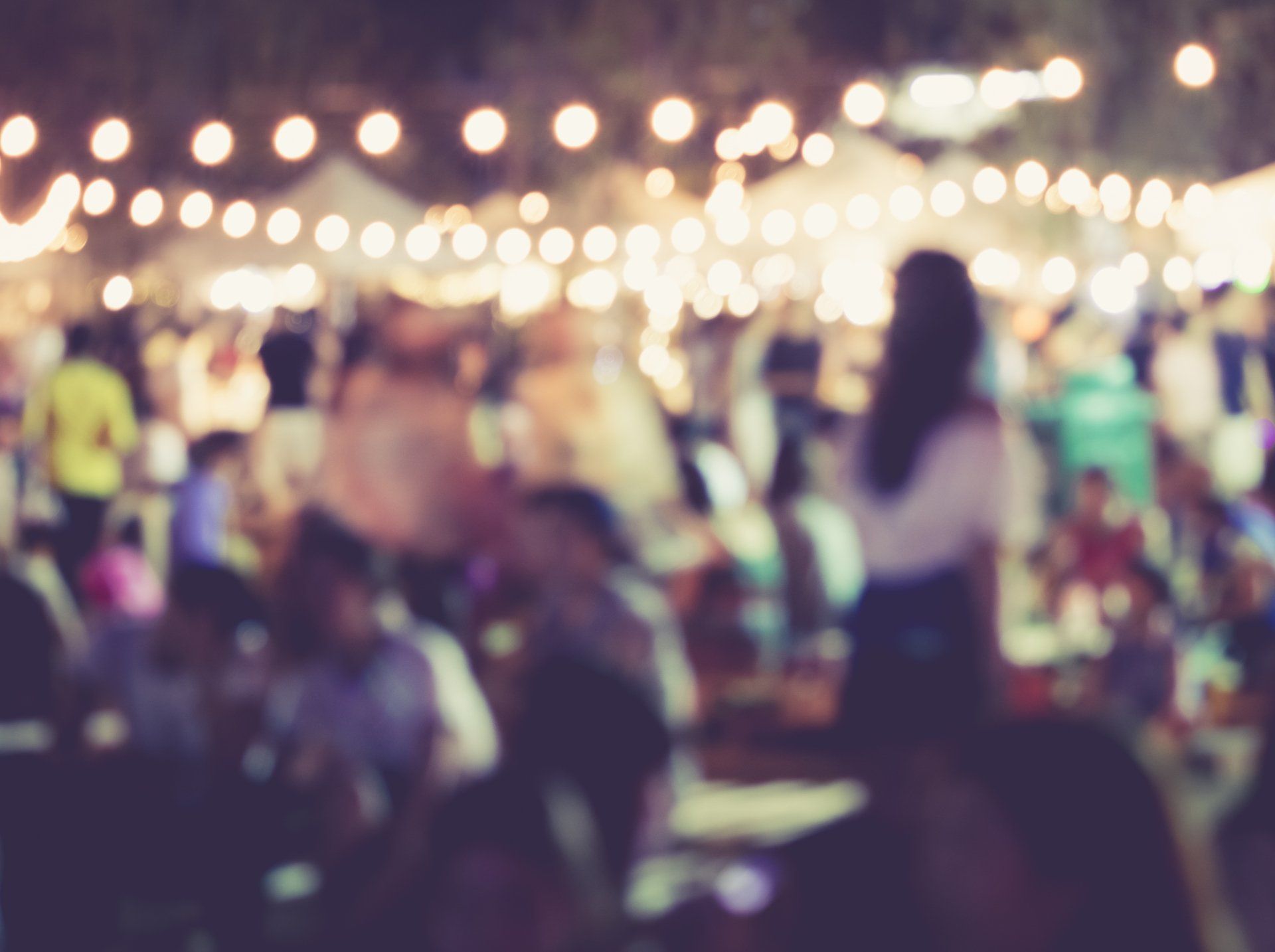 Blurred image of a crowded outdoor event at night with string lights and people socializing.