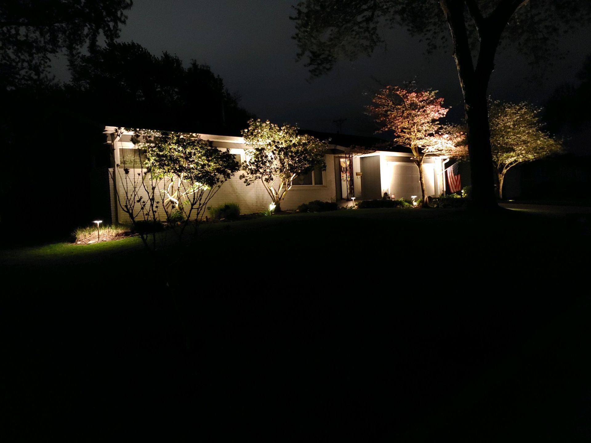 House illuminated at night by landscape lighting, with trees and shrubs, on a grassy lawn.