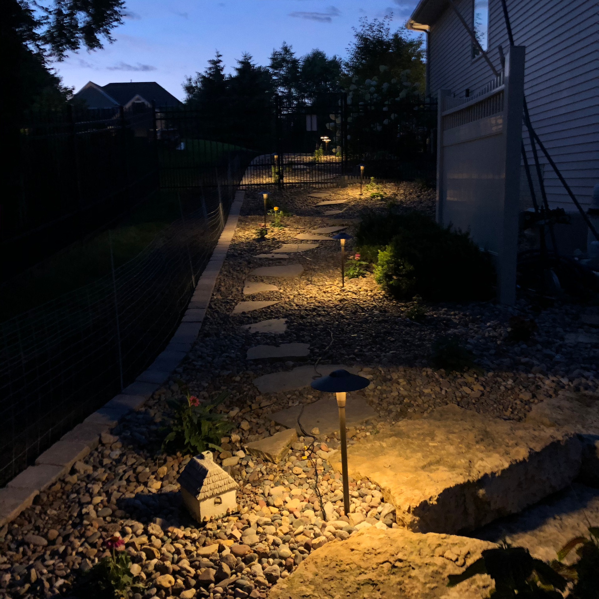 Gravel pathway with lit landscape lights, leading uphill past a house and retaining wall at dusk.