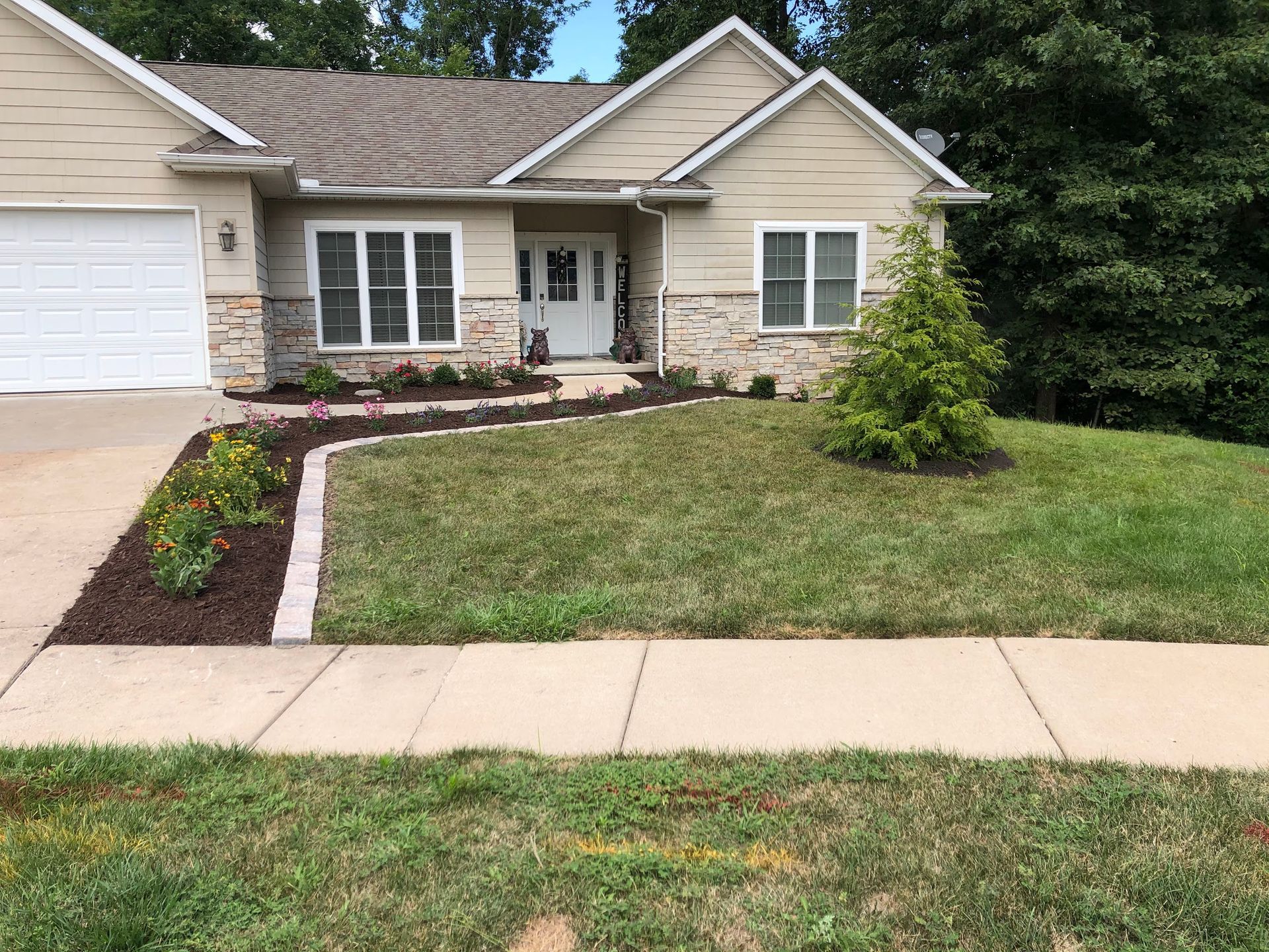Tan house with stone accents, well-kept lawn, flowerbeds with colorful blooms, sidewalk.