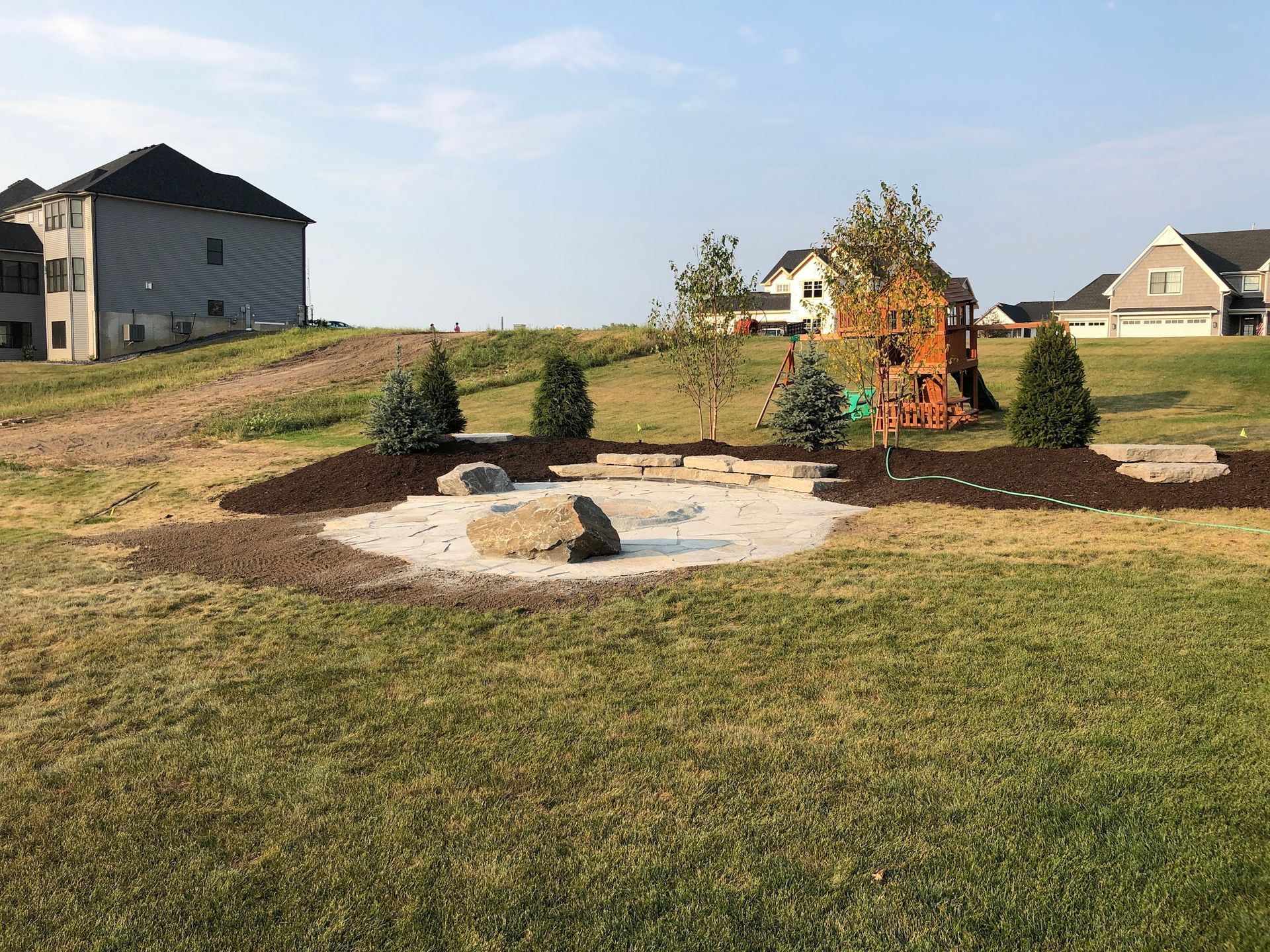 Backyard fire pit surrounded by grass, landscaping, and houses in the background.