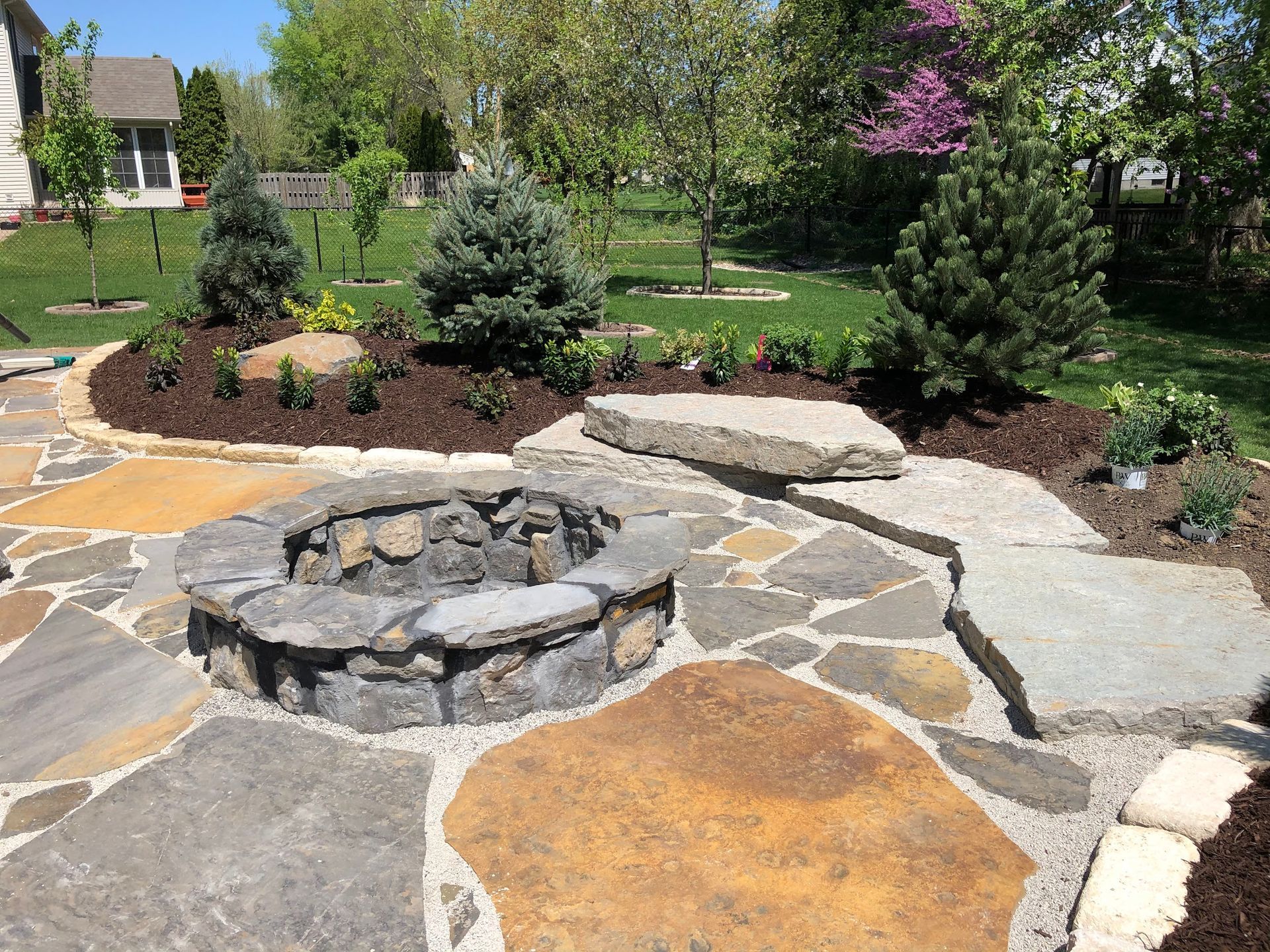 Stone patio with fire pit, landscaping with evergreens, and a grassy backyard.