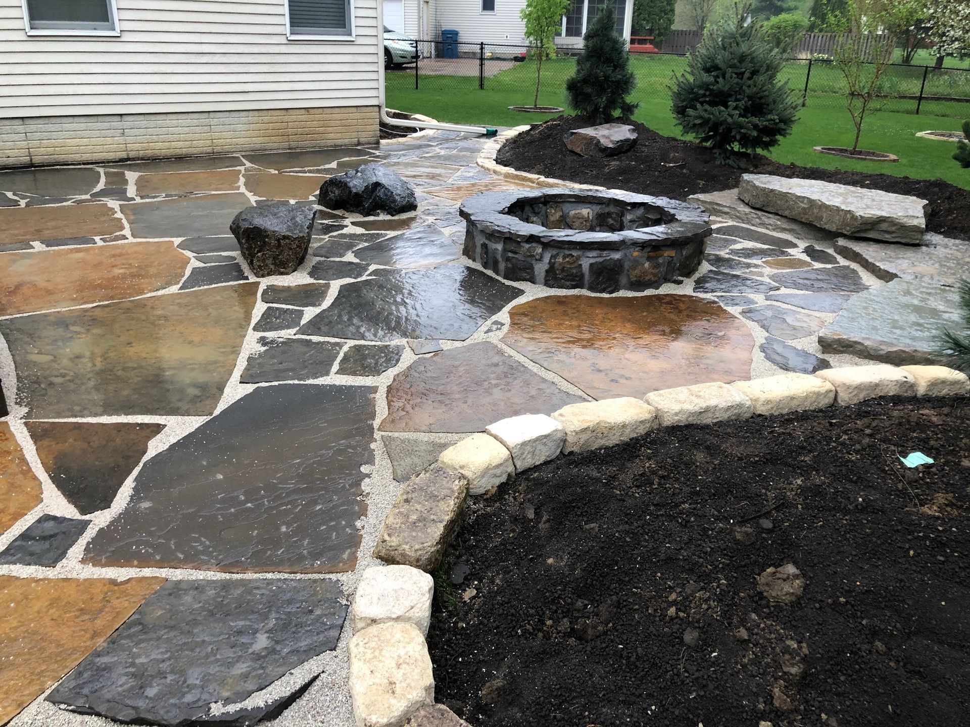 Stone patio with fire pit, boulders, and garden bed next to a house.