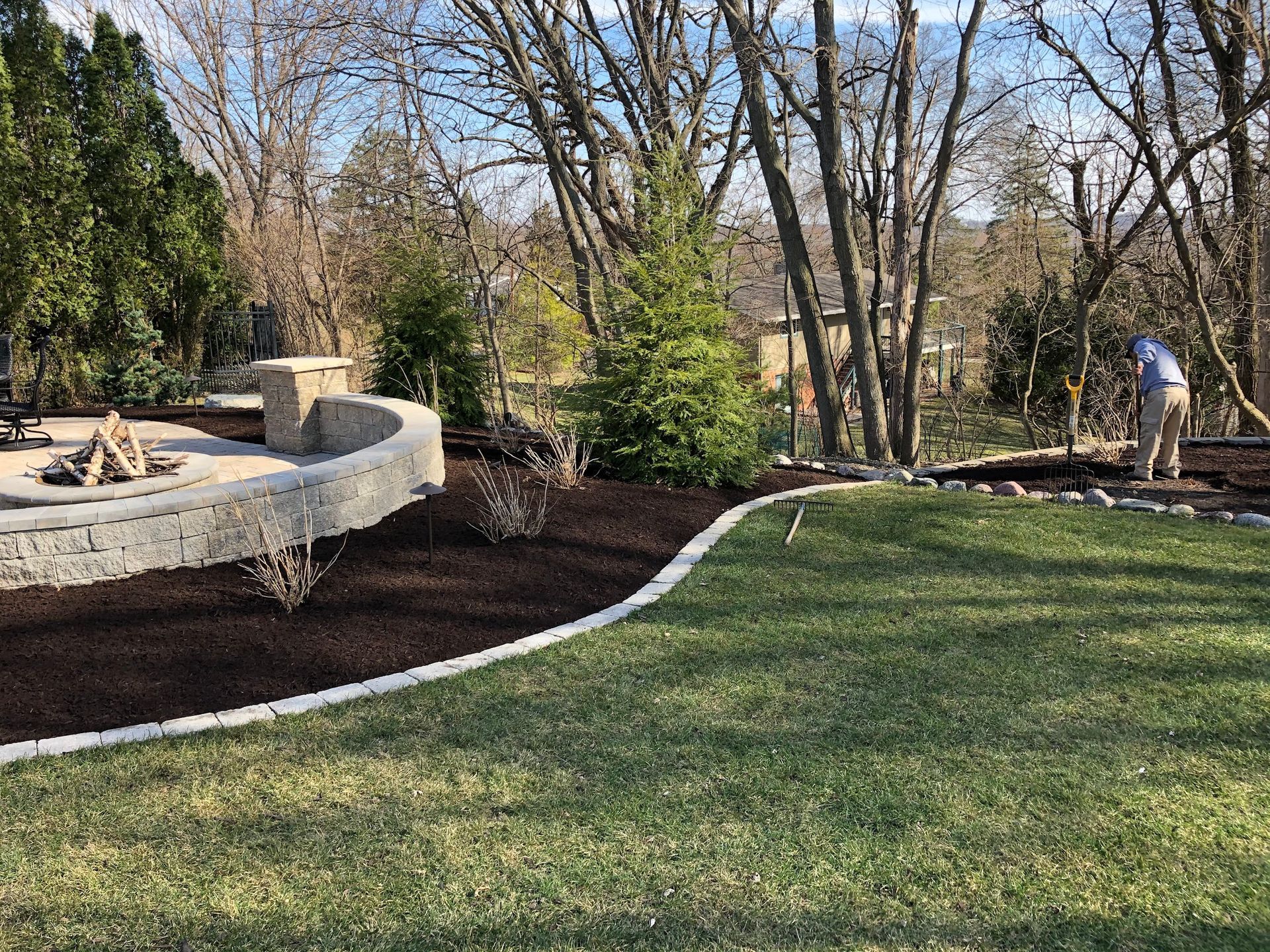 A person mulches a garden bed with dark mulch. Lawn in the foreground. Firepit to the left.