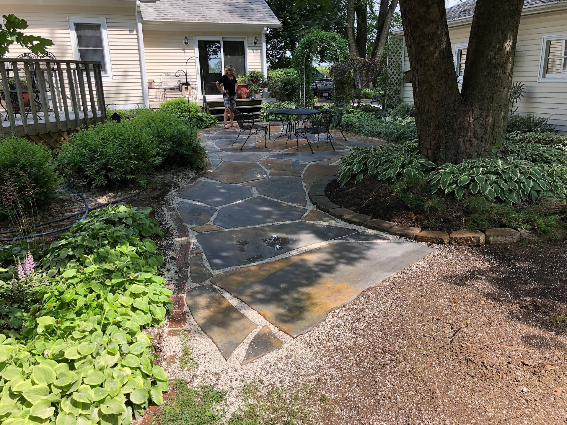 Flagstone patio with a table, surrounded by greenery, next to a house.