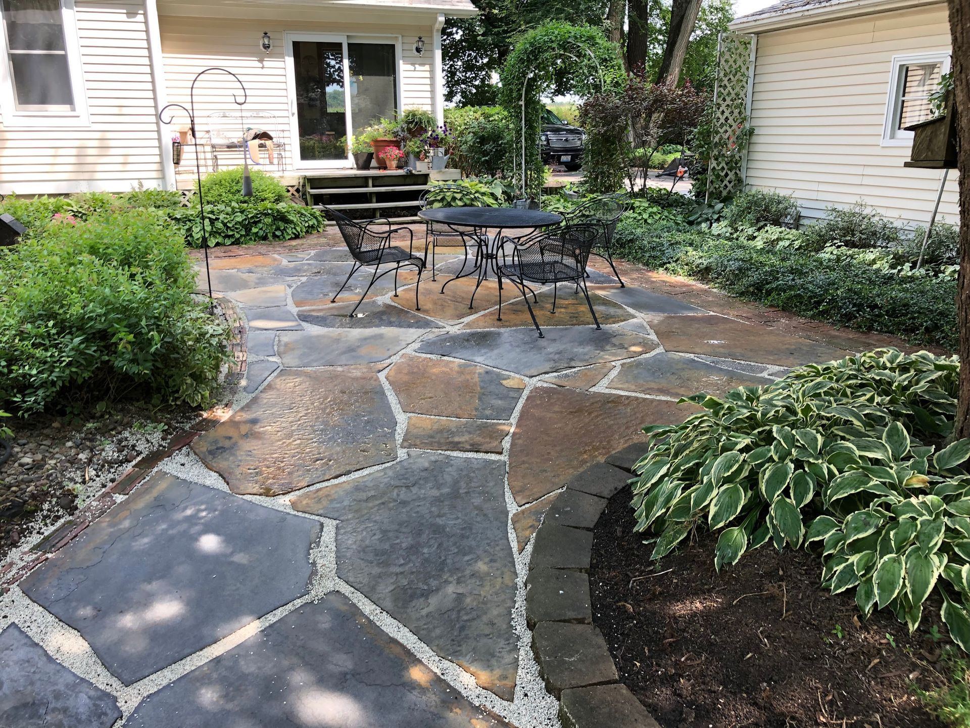 Stone patio with table and chairs surrounded by greenery, near a house.
