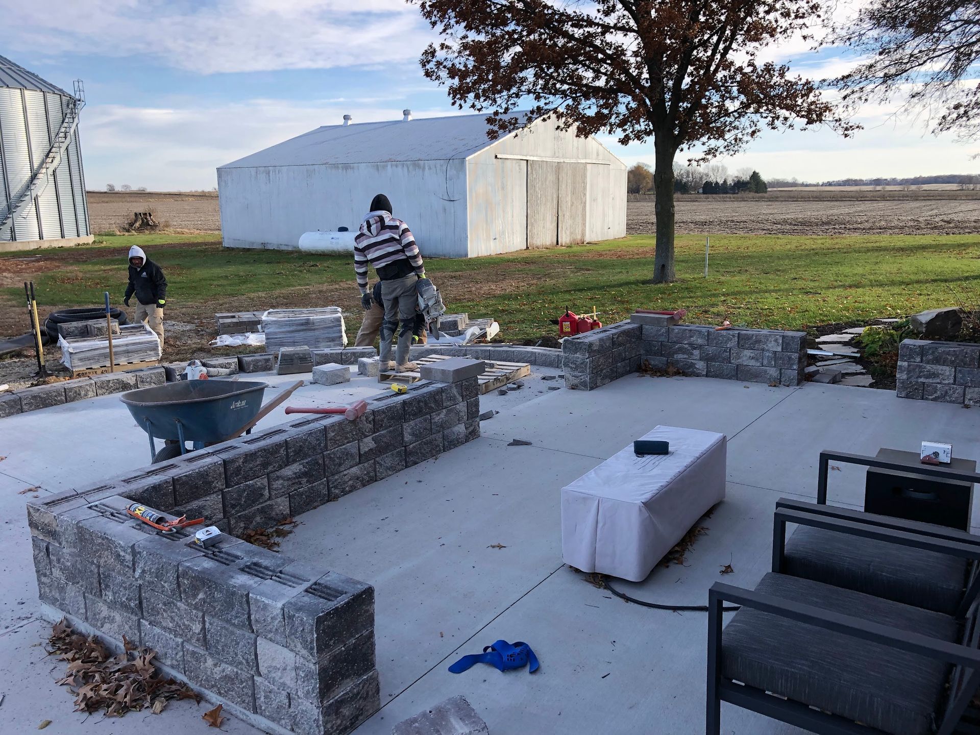 Construction workers building a stone patio. Gray blocks, concrete base, rural setting with a barn.