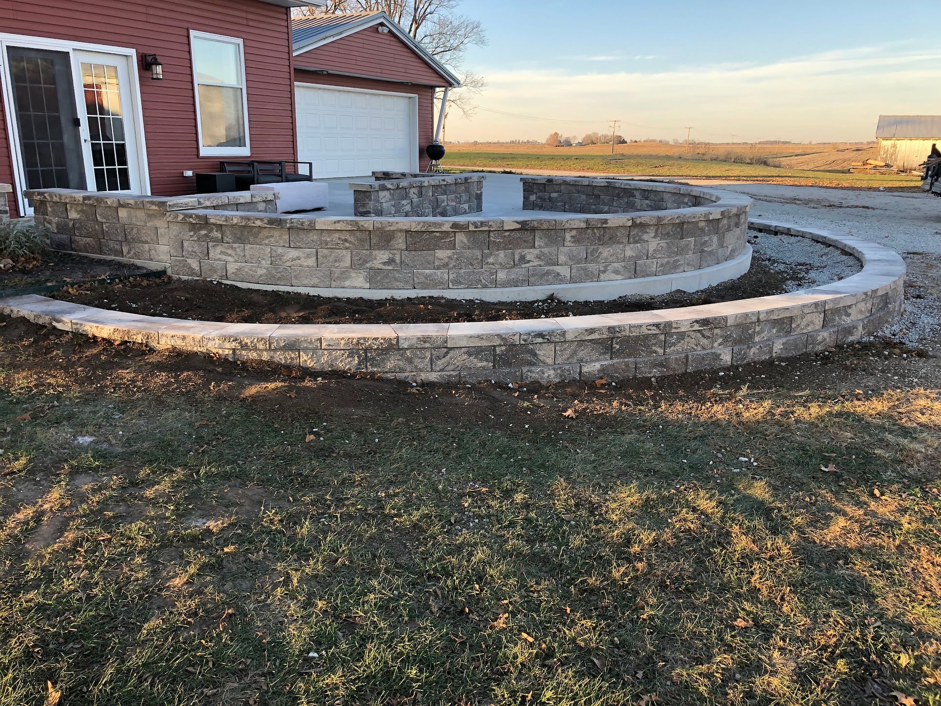 Circular stone patio with tiered walls in a grassy yard, red house and white garage in background.