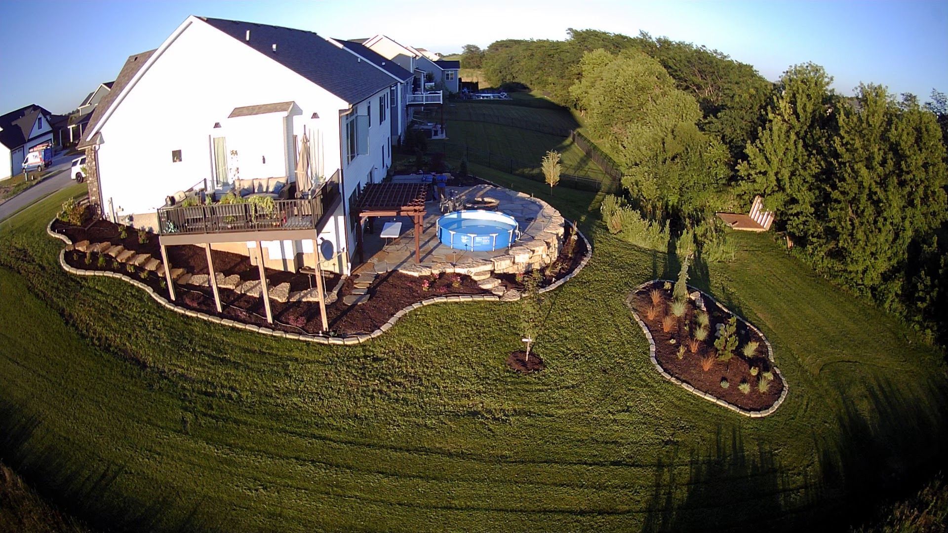 Aerial view of a backyard with a pool, deck, and landscaping; green grass and a large white house.